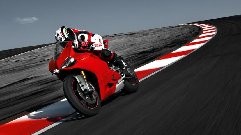 A red Ducati sportbike leans into a curve on a desaturated racetrack with red and white curbing and motion blur.