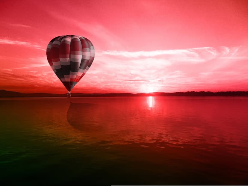 Hot air balloon floating above a vibrant red lake at sunset with a mirrored reflection on the calm water surface.