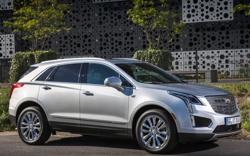 Silver Cadillac XT5 SUV parked in front of a dark grey perforated building with green trees in daylight.