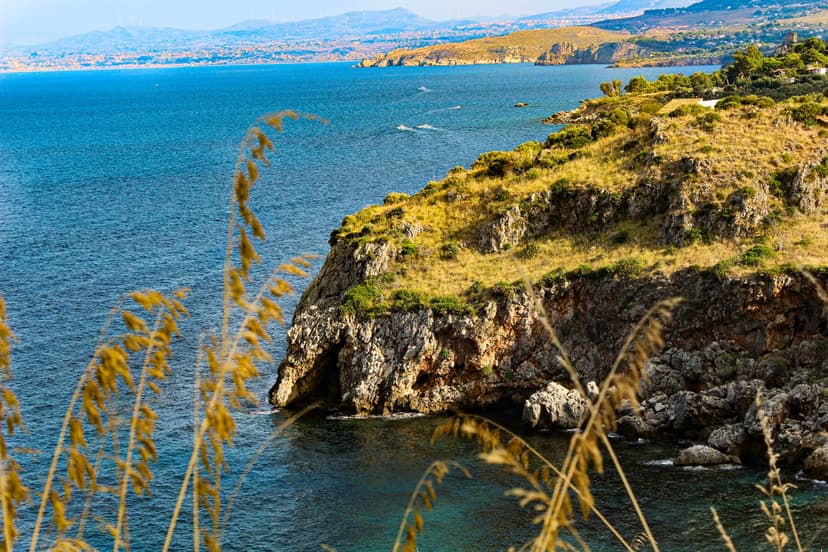 Rugged Sicilian cliffs with green shrubs overlook deep blue sea water under a hazy horizon with golden grass.
