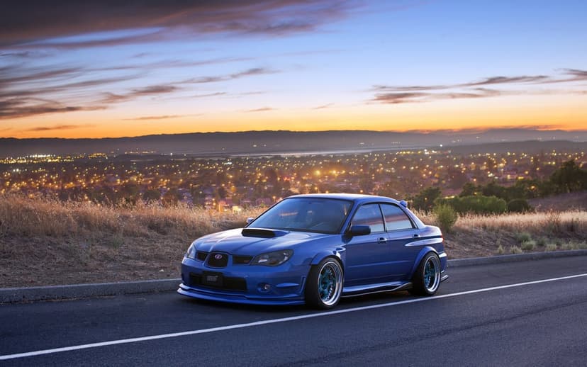 Blue Subaru WRX STI parked on a hillside road overlooking a city at sunset with orange and purple clouds.