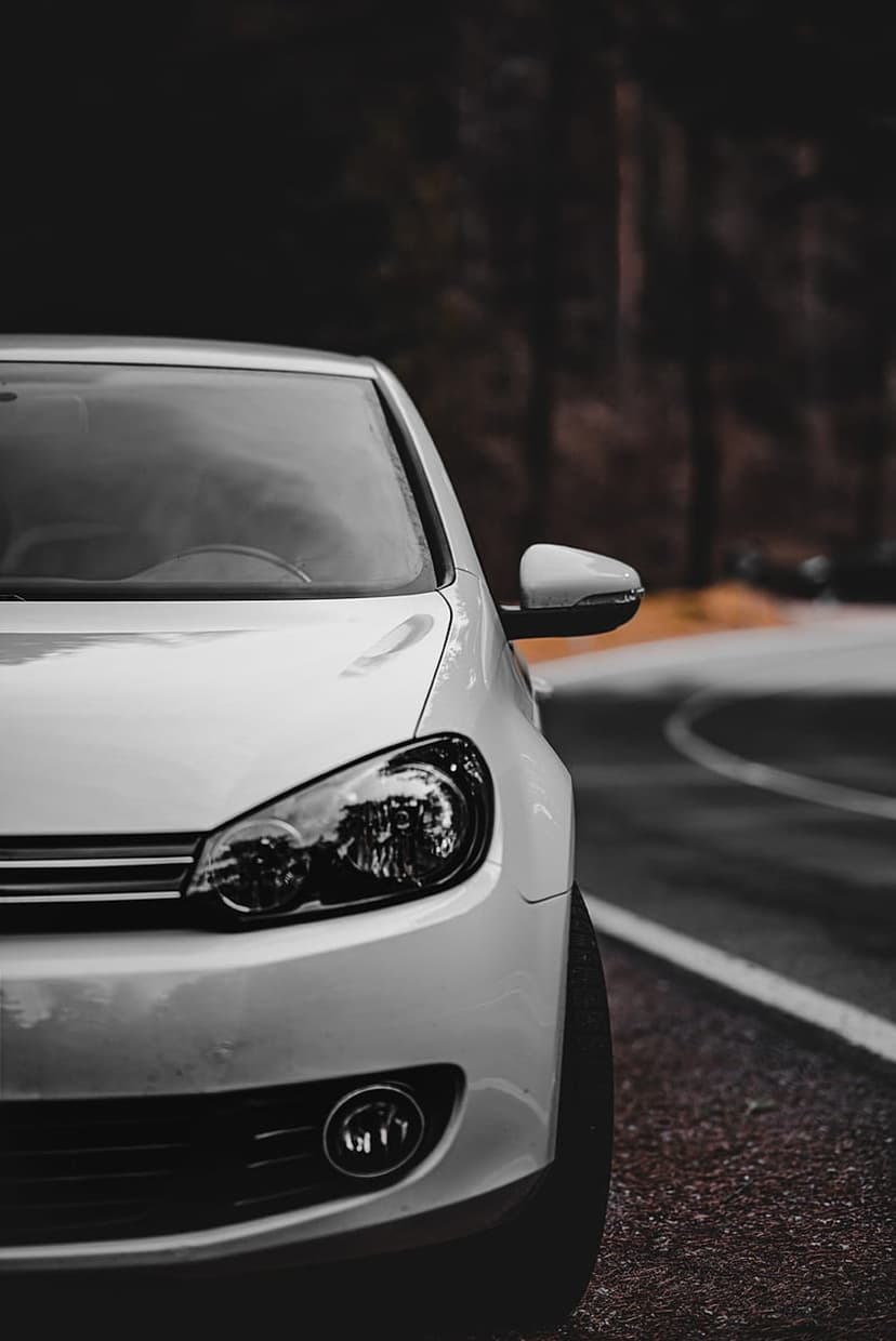 White car headlight and side mirror reflect dim light on a curved asphalt road through a dark pine forest.