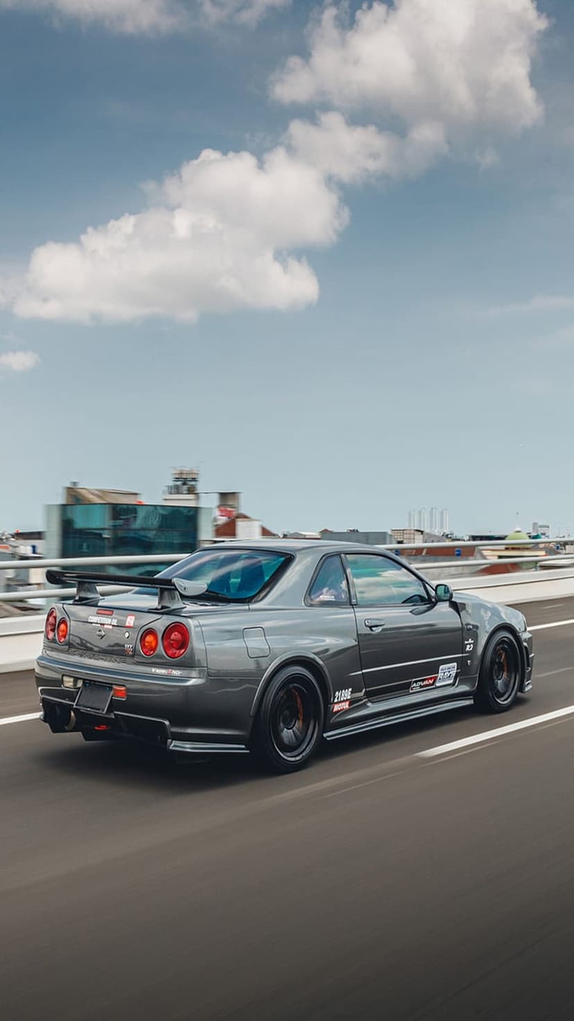 Nissan Skyline R34 GT-R with a large spoiler and round taillights driving on a blurred highway under clouds.