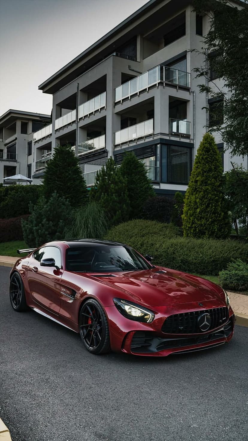 Red Mercedes-AMG GT S Coupe parked on a suburban road beside a modern house with manicured green hedges.