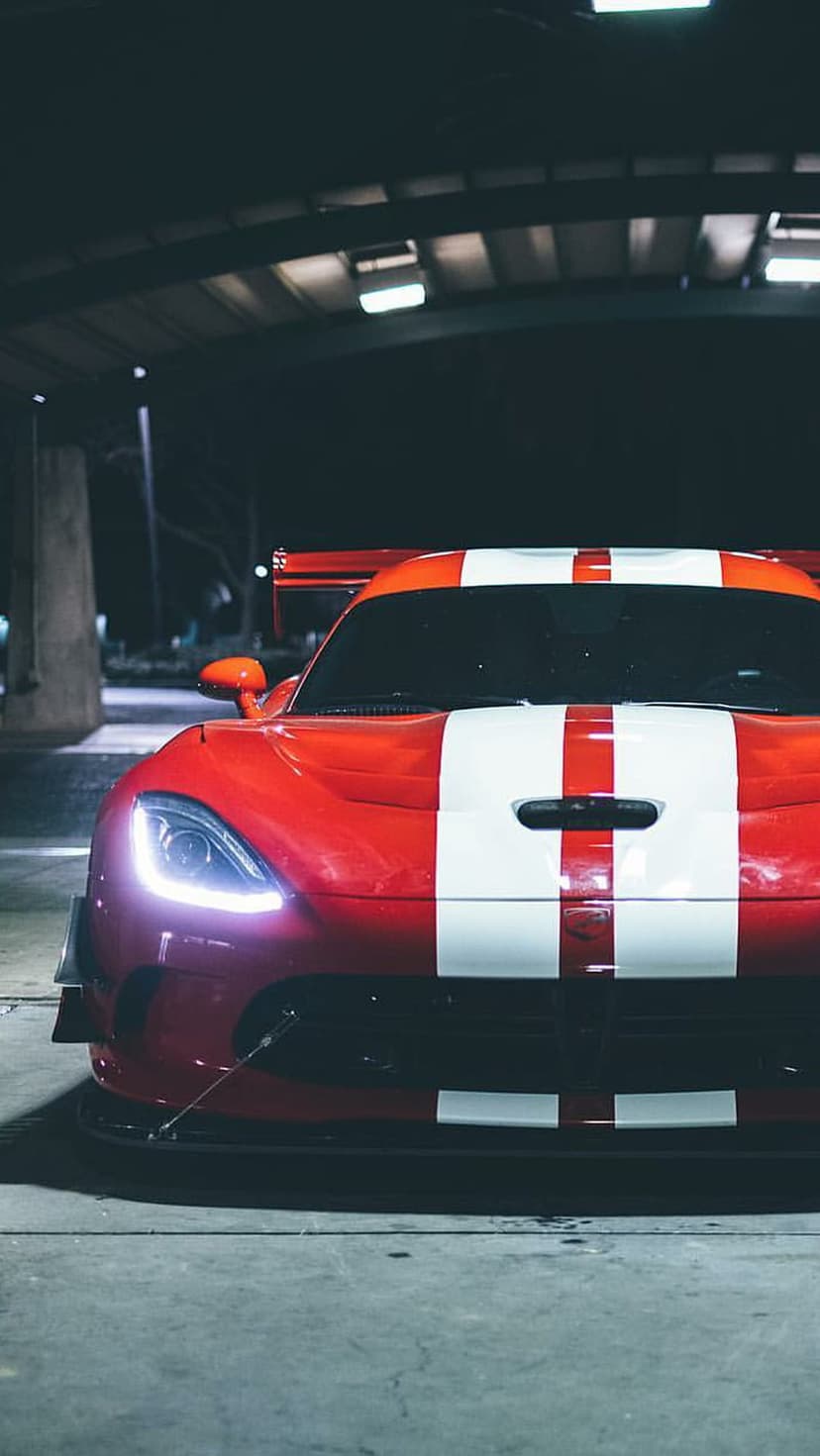 Red Dodge Viper with white racing stripes and glowing headlights parked under a dark structure at night.