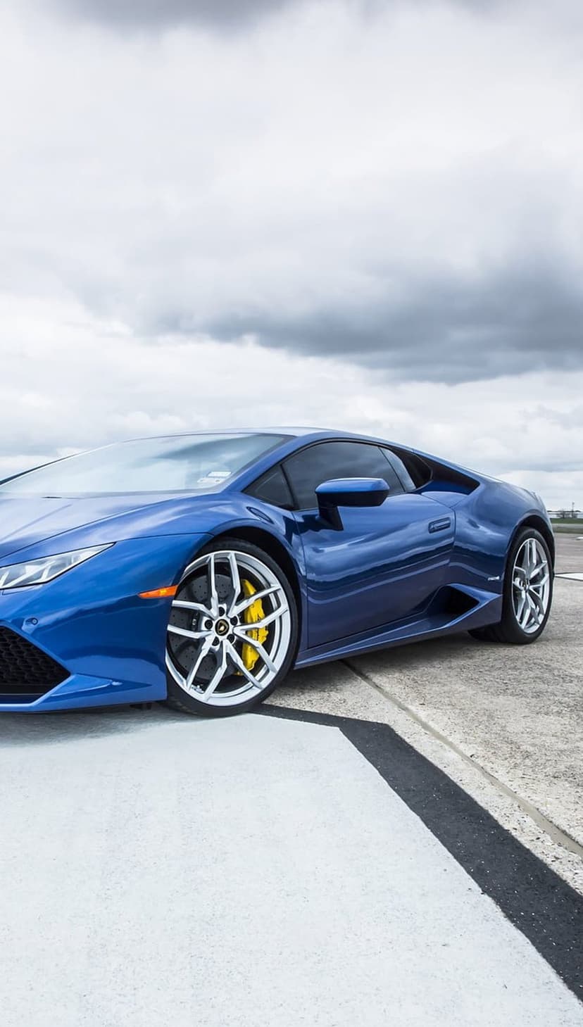 Blue Lamborghini Huracan supercar on gray concrete under a cloudy sky featuring visible yellow brake calipers.