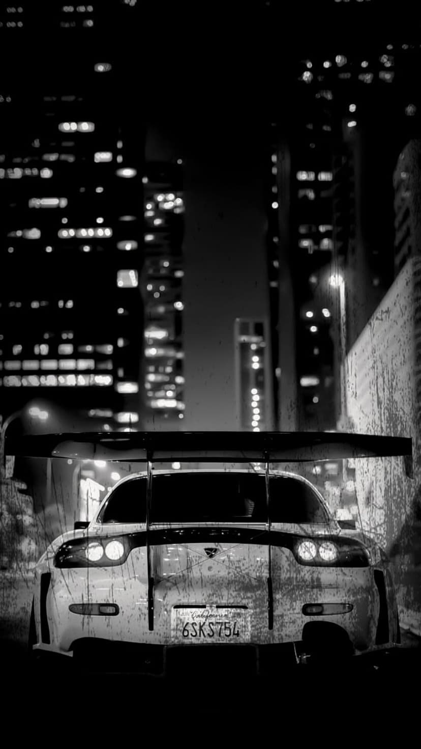 Mazda RX-7 with a large rear wing parked in a dark city alley featuring blurred urban lights and grainy texture.