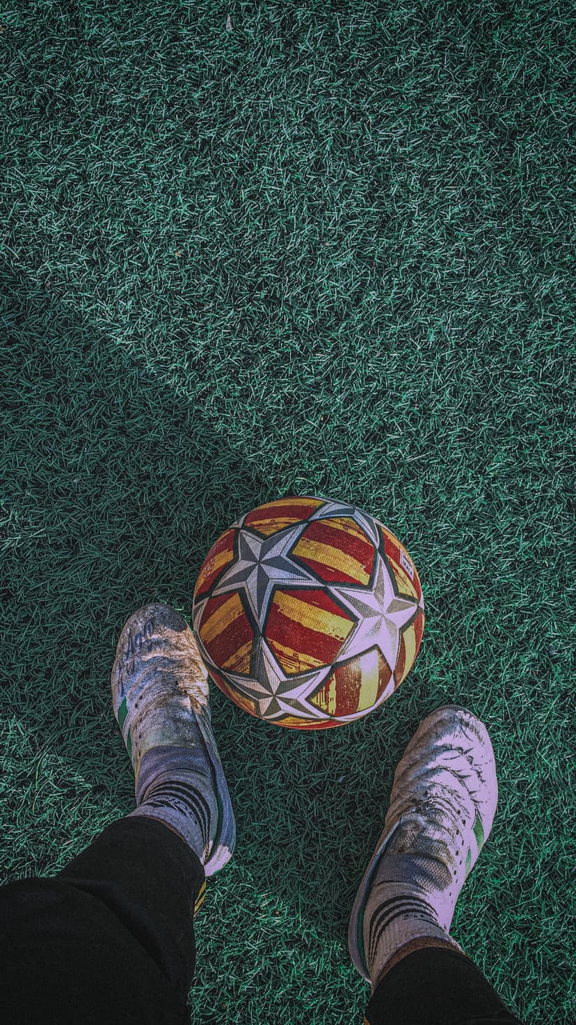 Star-patterned soccer ball between two feet wearing athletic cleats on a green artificial turf sports field.