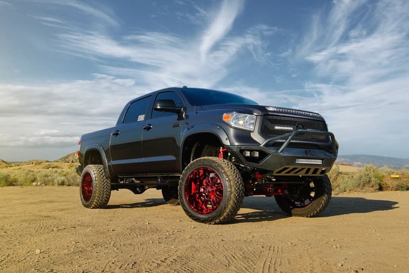 Lifted dark grey pickup truck with metallic red wheels parked on a desert dirt path under a wispy blue sky.