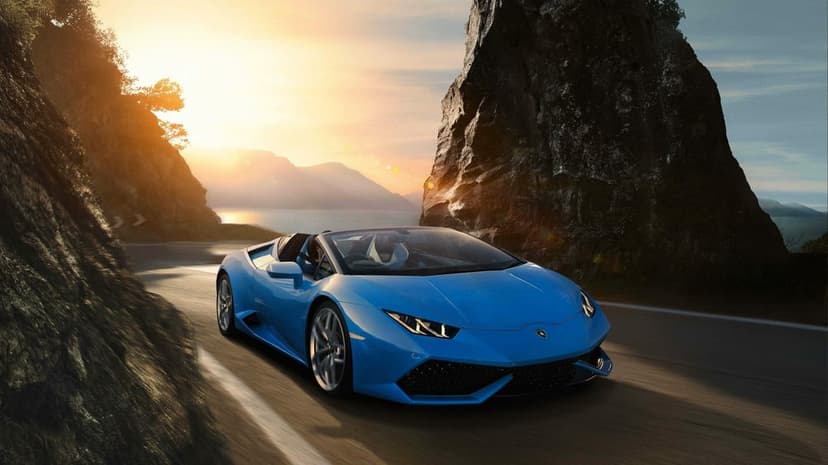 Blue Lamborghini Huracán Spyder driving fast on a winding mountain road at sunset between rocky cliffs.