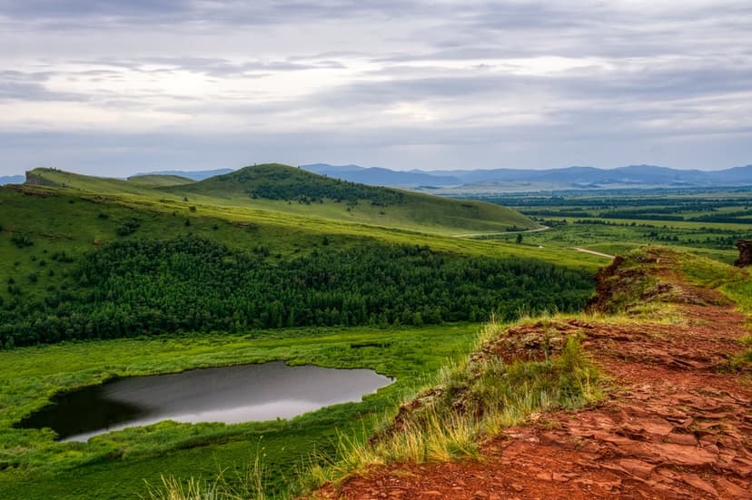 Rolling green hills and a pine forest surround a calm lake under a cloudy sky viewed from a rocky ledge.