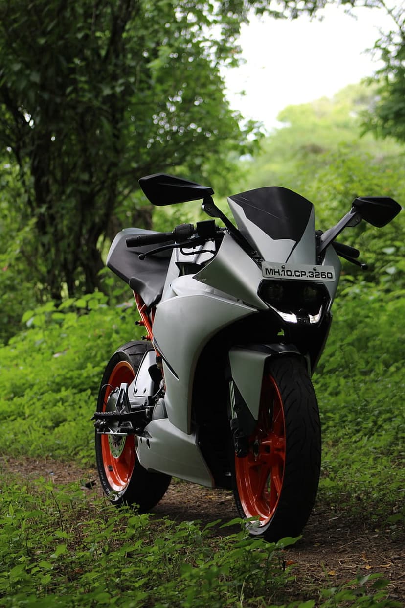 KTM RC 390 sportbike with orange wheels parked on a dirt trail surrounded by dense green forest foliage.