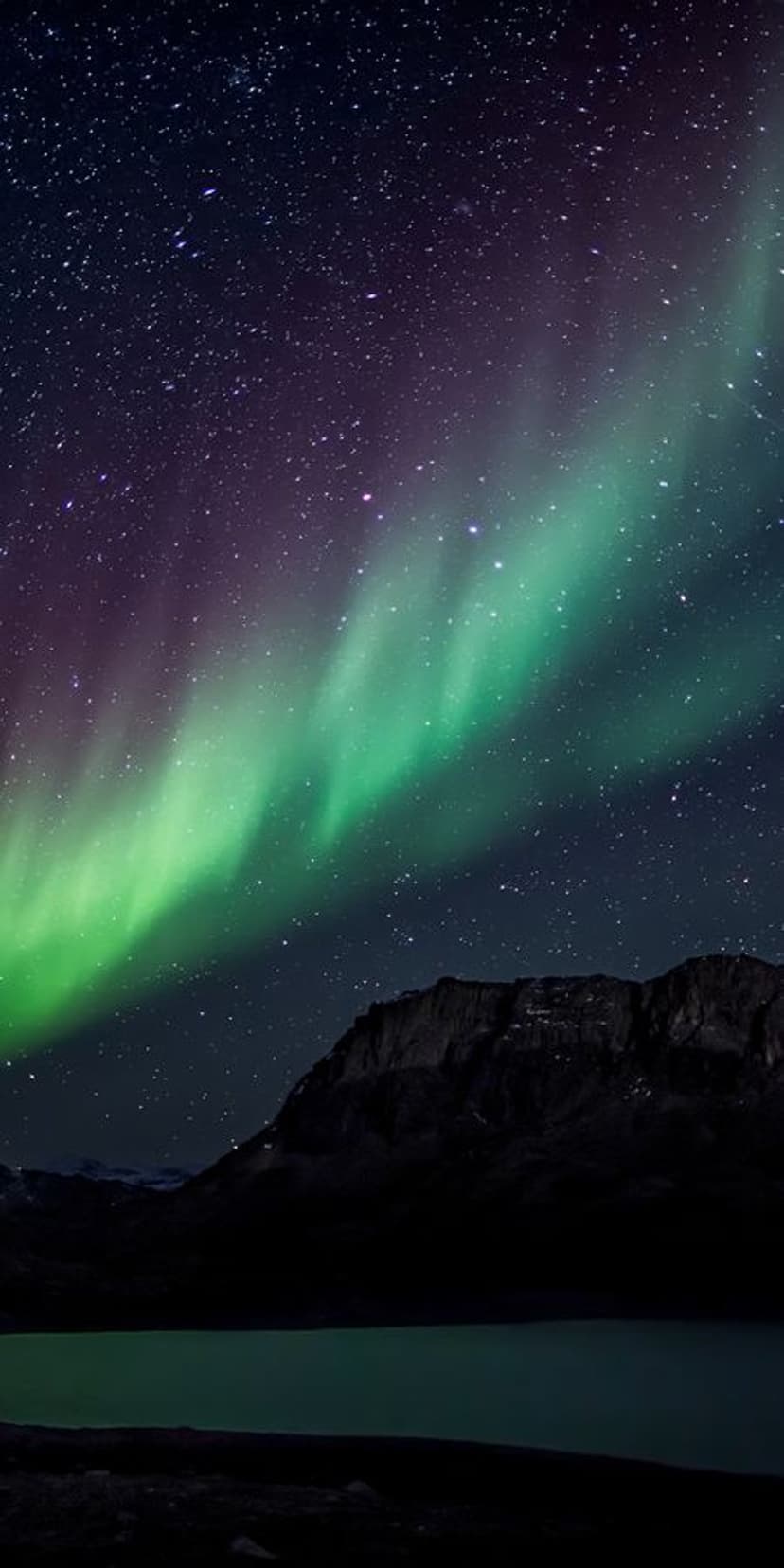 Green aurora borealis ribbons stretch over jagged mountain peaks reflecting in a calm dark lake at night.