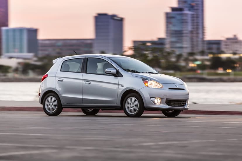 Silver Mitsubishi Mirage hatchback driving on a coastal road with a blurred city skyline and sunset sky.