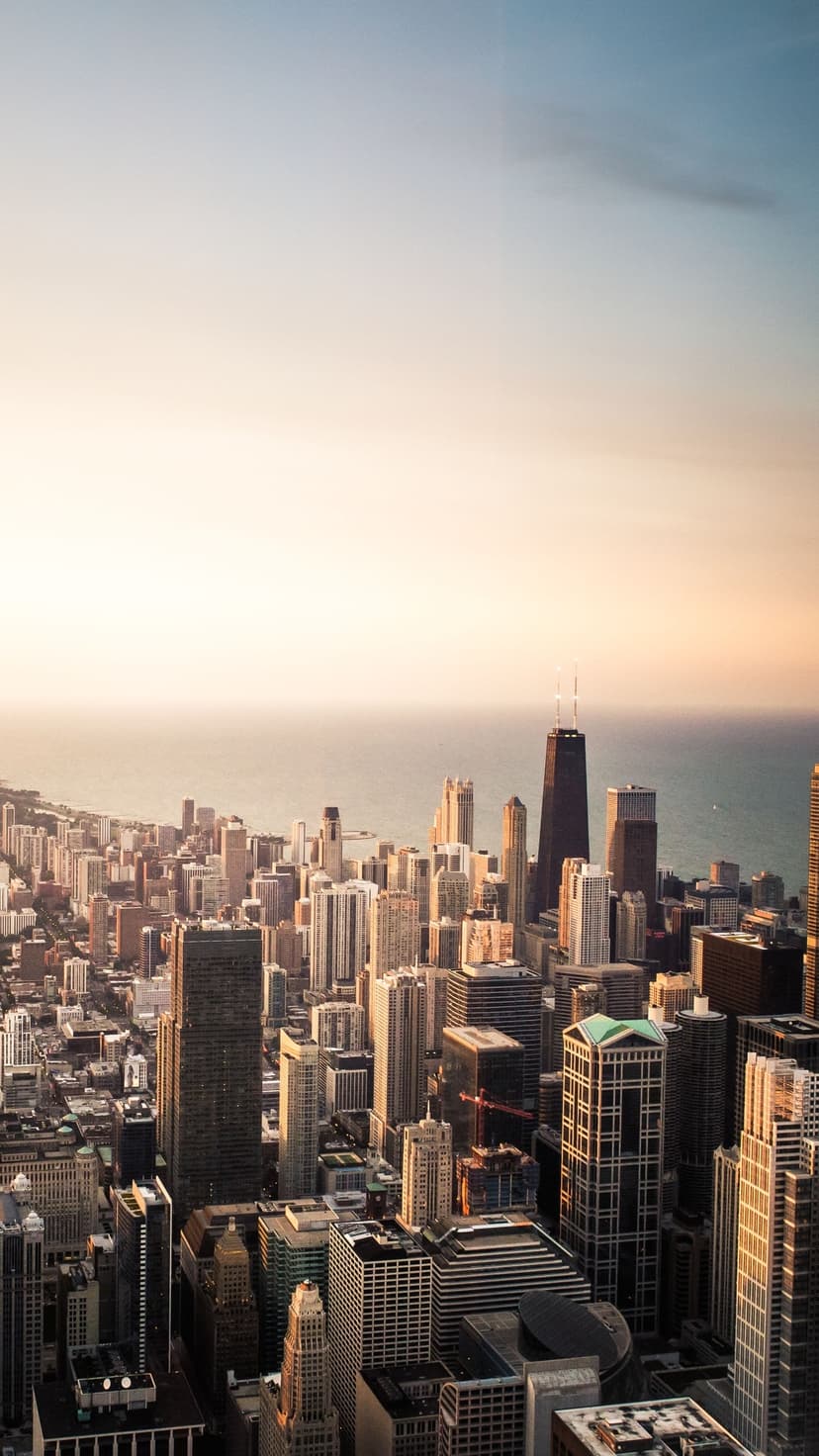 Skyscrapers and urban high-rises stand beside a vast lake under a golden sunset in an aerial perspective.
