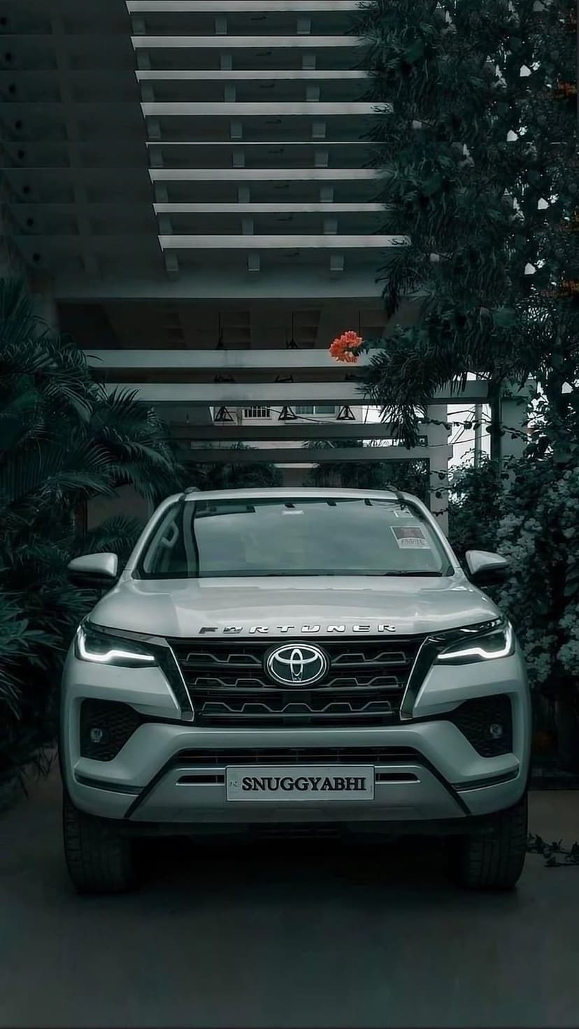 Toyota Fortuner SUV with glowing LED headlights parked near a modern building and lush green plants.