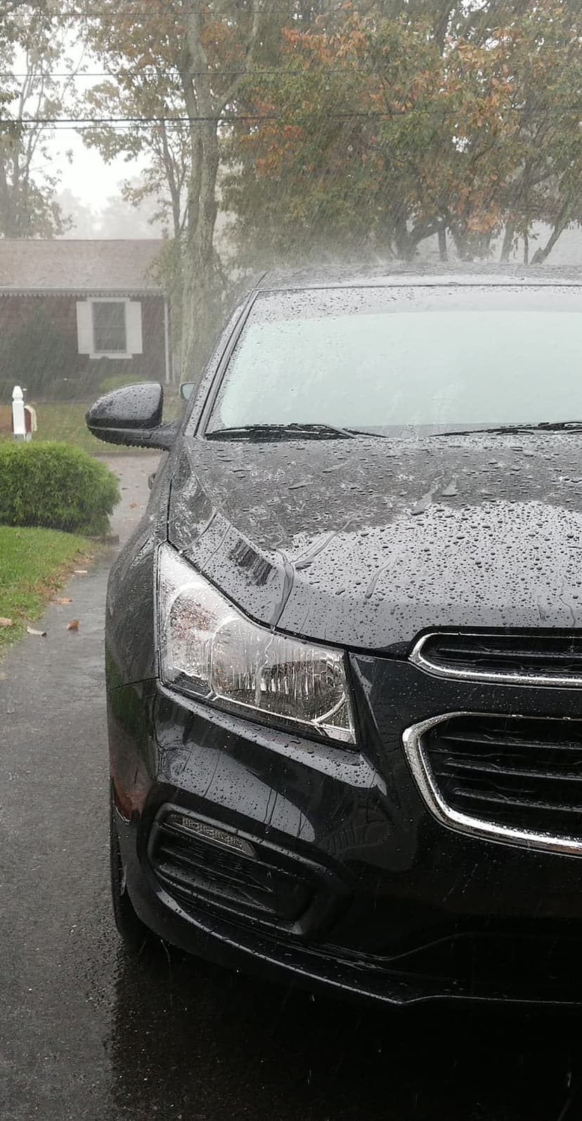 Black car hood covered in raindrops reflecting an overcast sky with visible headlights and front grille.