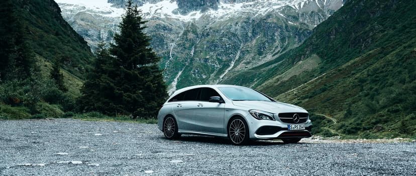 Silver Mercedes-Benz station wagon on a gravel road with steep rocky mountains and green trees in the background.
