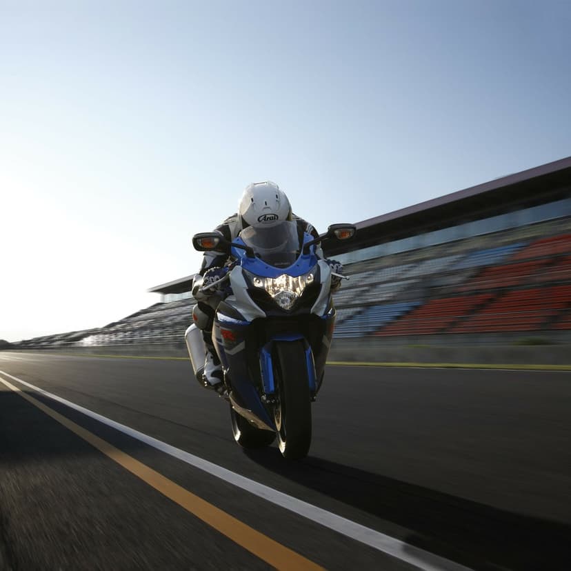 Blue and white Suzuki GSX-R motorcycle racing on a track past blurred grandstands under a clear sky.