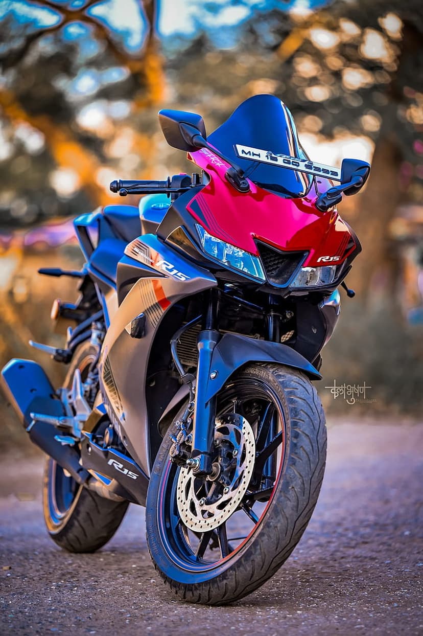 Yamaha R15 V3 motorcycle in red and blue with sharp headlights and alloy wheels against a blurred green backdrop.