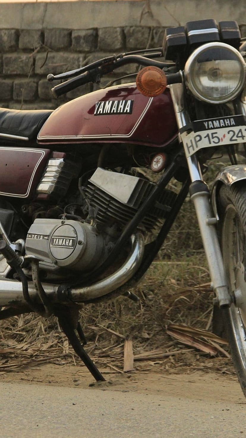 Maroon Yamaha RD350 motorcycle with chrome details parked on a dirt path next to a weathered brick wall.