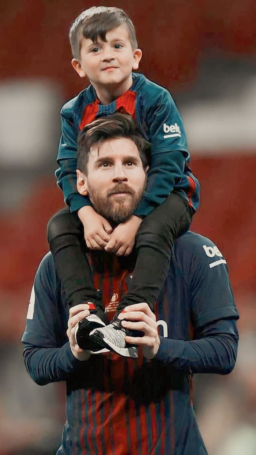 Lionel Messi carries his son Thiago on his shoulders while wearing matching Barcelona kits in a crowded stadium.