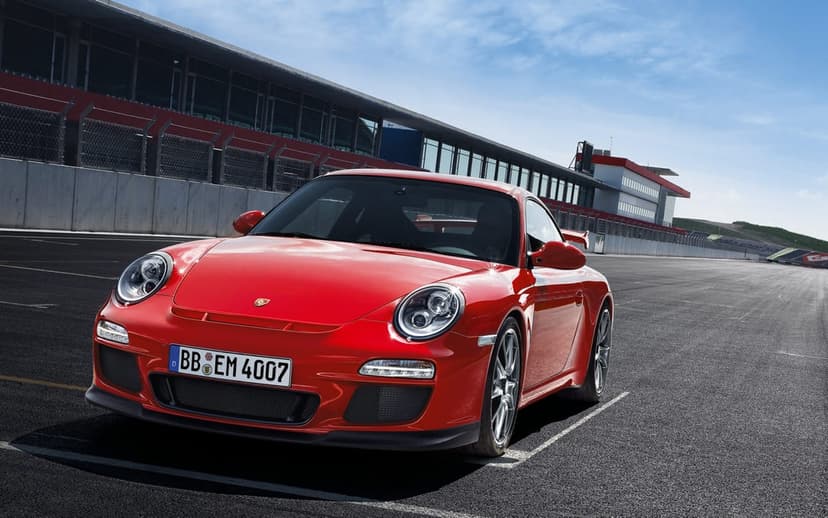 Red Porsche 911 GT3 parked on an asphalt racetrack with grandstands and a bright blue sky background.