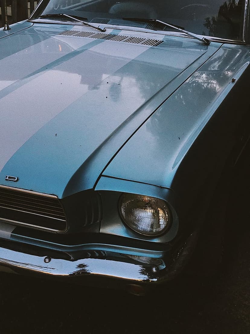 Blue vintage car hood with white racing stripes, chrome bumper, and round headlight in a close-up view.