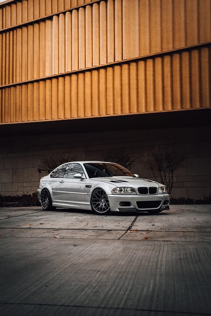 Silver BMW M3 E46 coupe parked on concrete near a modern building featuring vertical warm wooden panels.