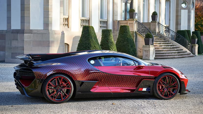Side view of a red and black Bugatti Divo supercar with diamond patterns parked on gravel by a stone mansion.