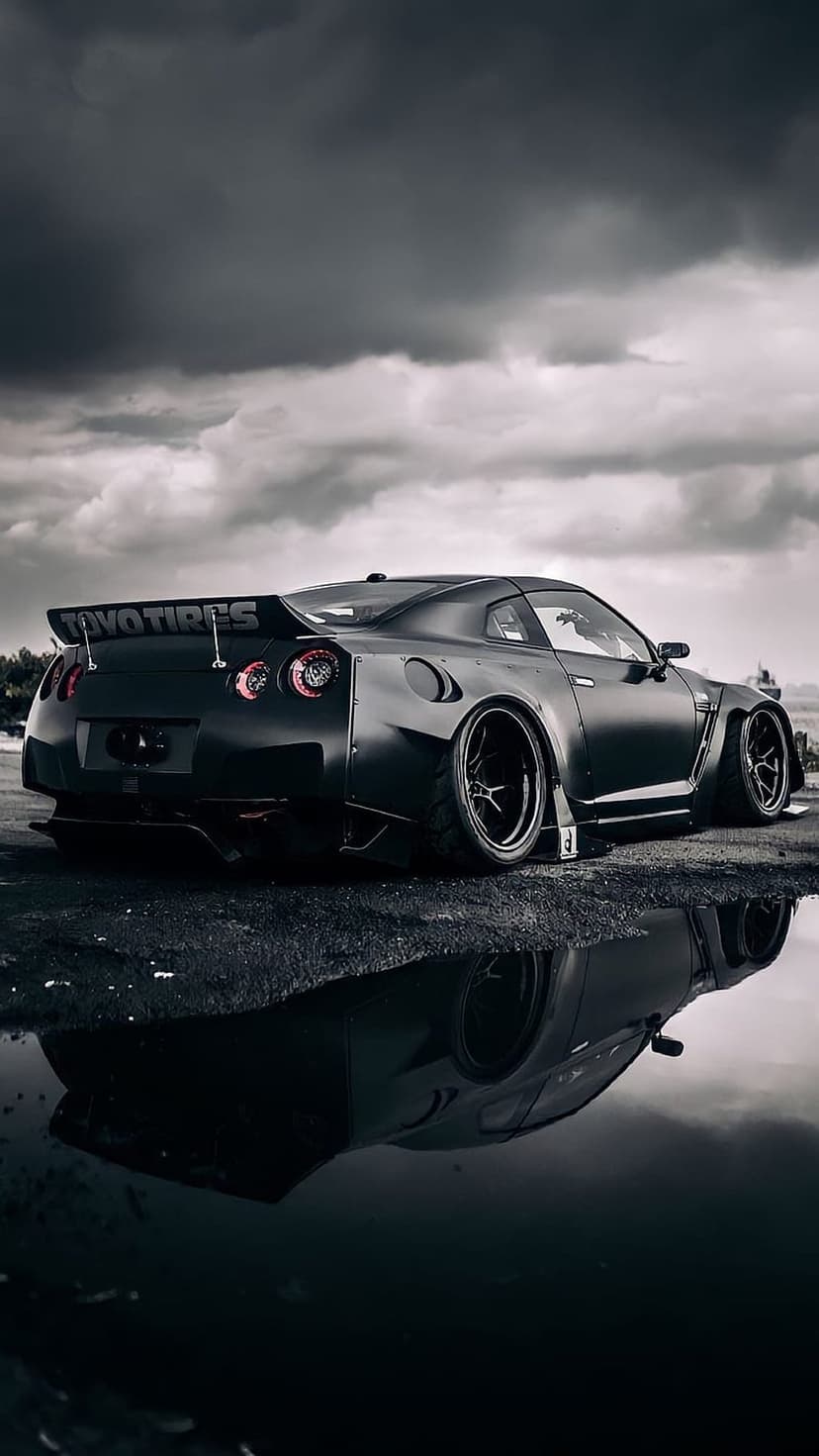 Nissan GT-R with a matte black widebody kit and rear wing reflects in a puddle under a cloudy, dark sky.