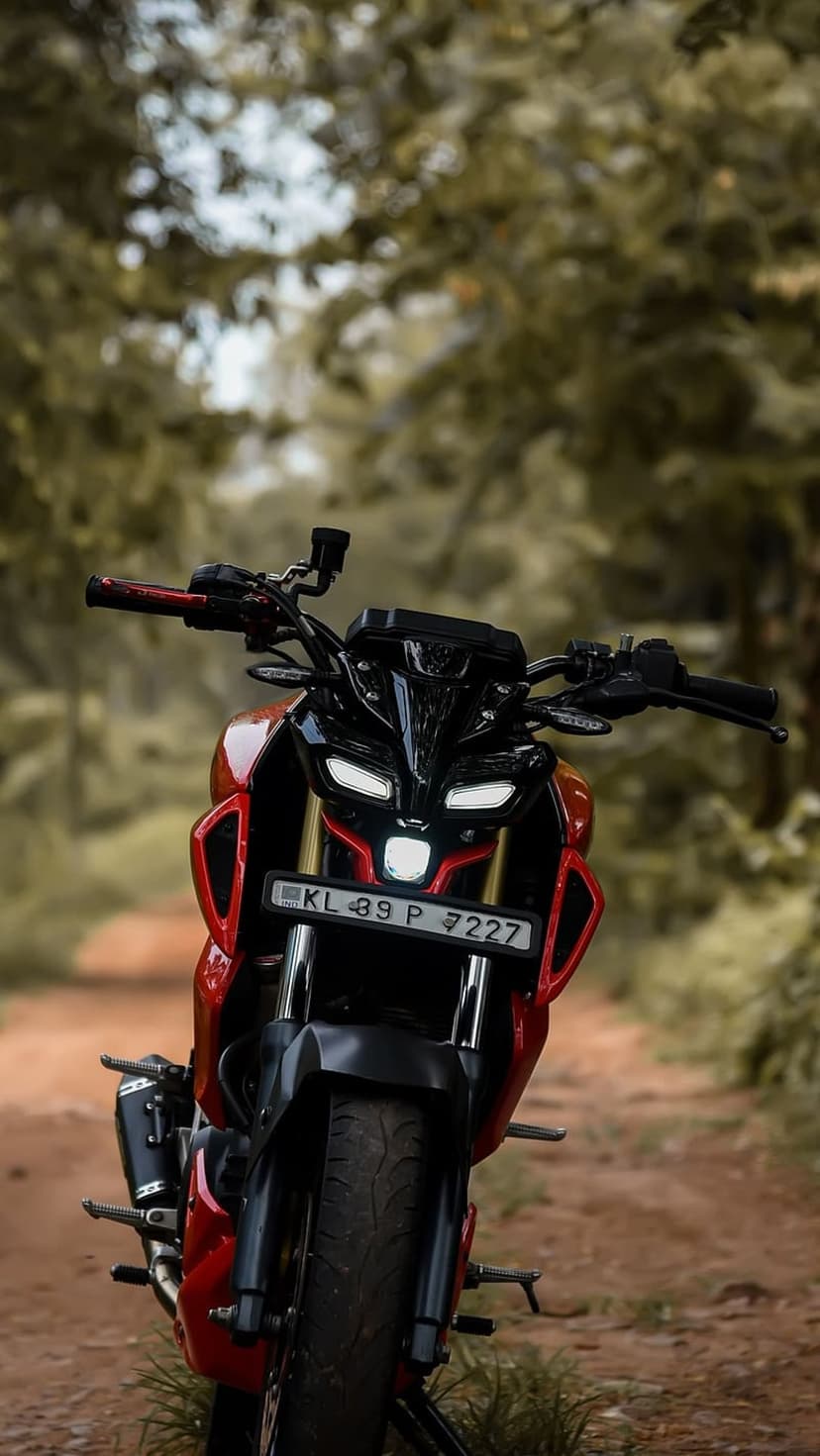 Red Yamaha MT-15 motorcycle with bright LED headlights parked on a dirt path surrounded by lush green trees.