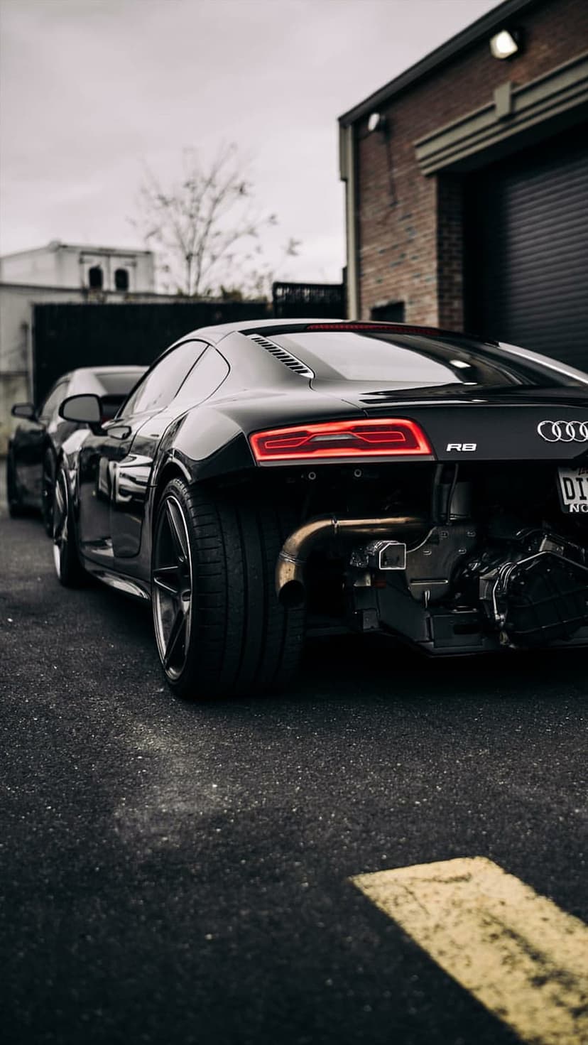 Audi R8 rear view showing red LED taillights and exposed engine parts on asphalt with industrial buildings.