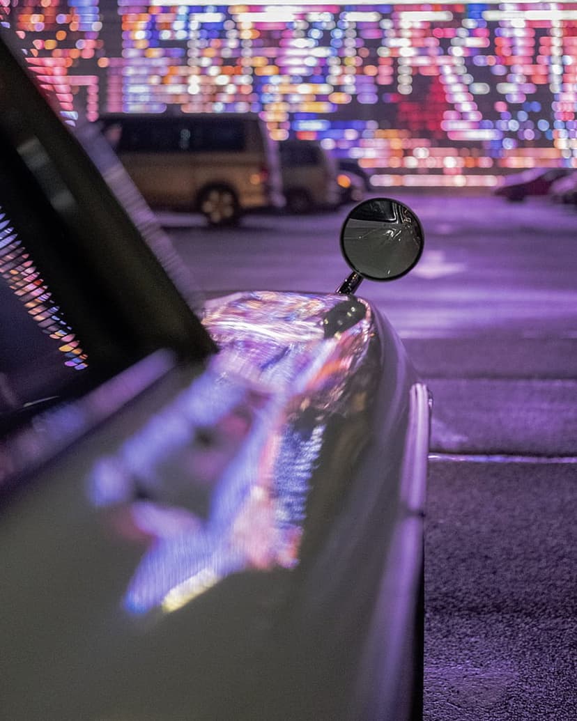 Car side mirror reflecting vibrant circular bokeh city lights on a dark metallic fender during a moody urban night.