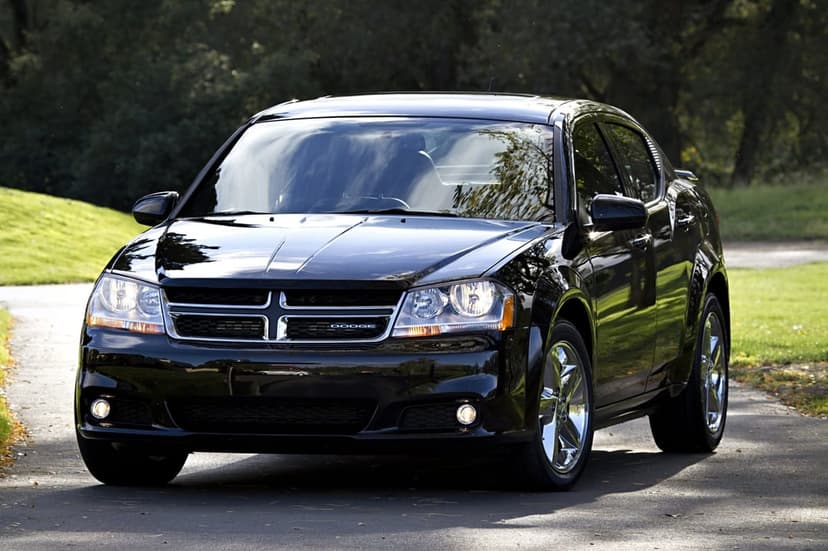 Black Dodge Avenger sedan parked on a paved road in a green park with chrome wheels and headlights on.