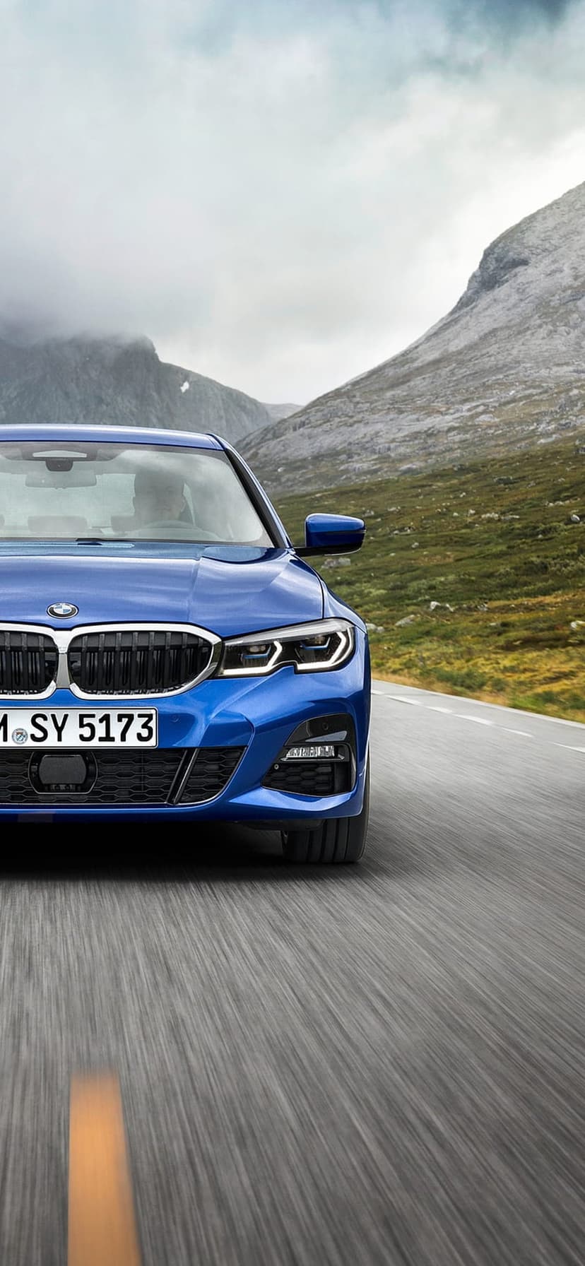 Blue BMW sedan driving on a winding asphalt road through green hills and rocky mountains under a cloudy sky.