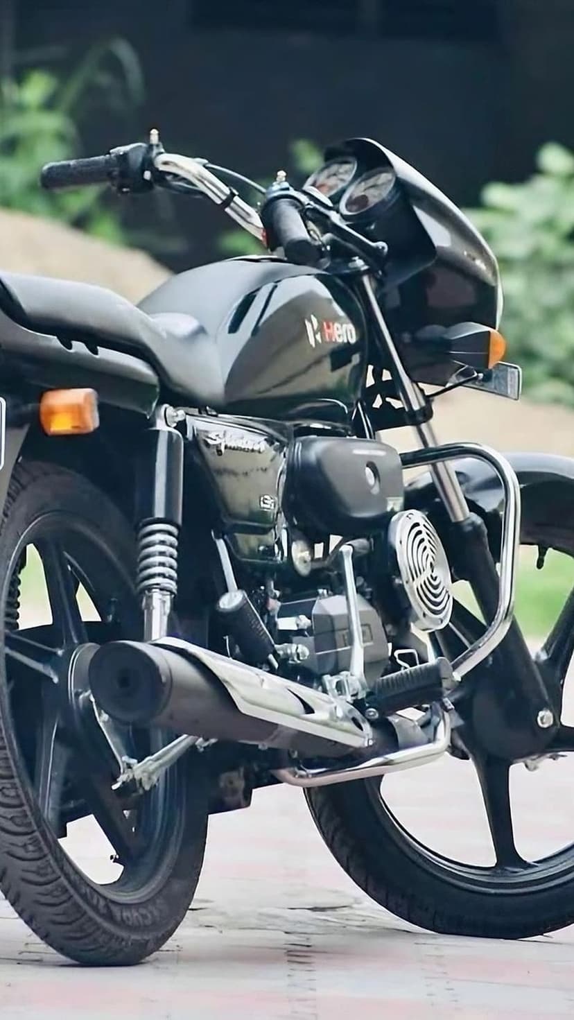 Black Hero Splendor motorcycle side profile showing the engine and exhaust against a blurred green background.