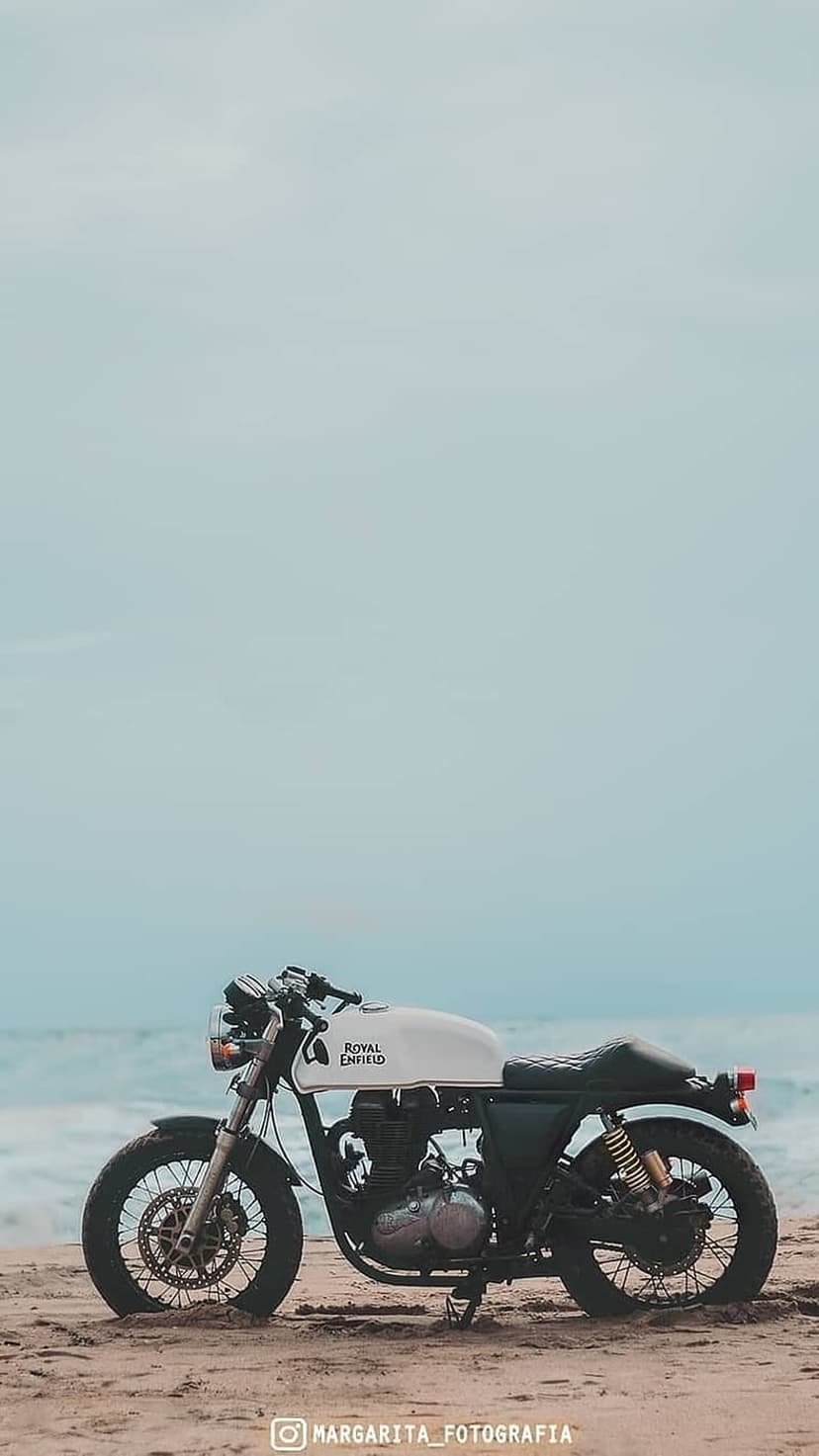 Royal Enfield motorcycle with a white tank and spoked wheels parked on a sandy beach near ocean waves.