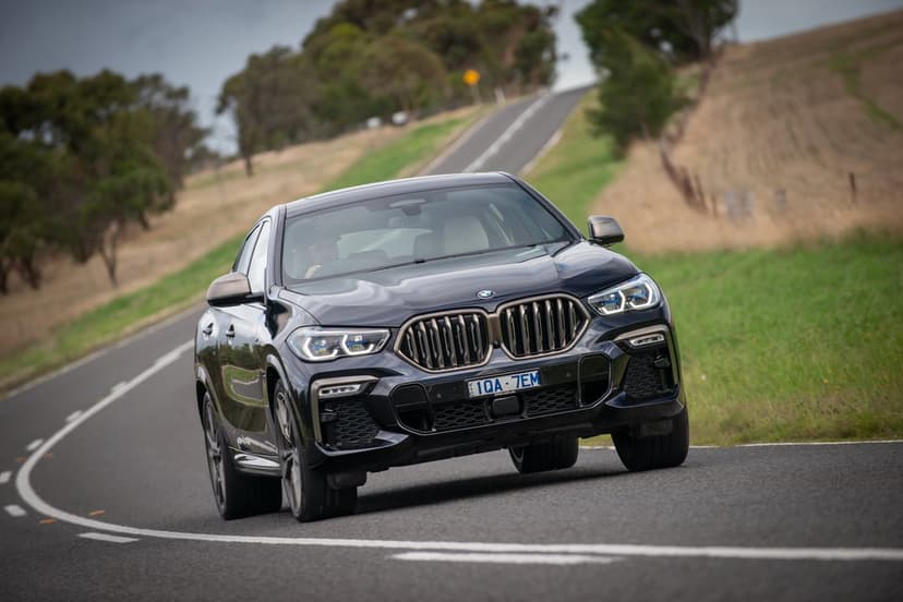 Dark blue BMW X6 M50i drives through a curved rural road surrounded by rolling green hills and dry fields.