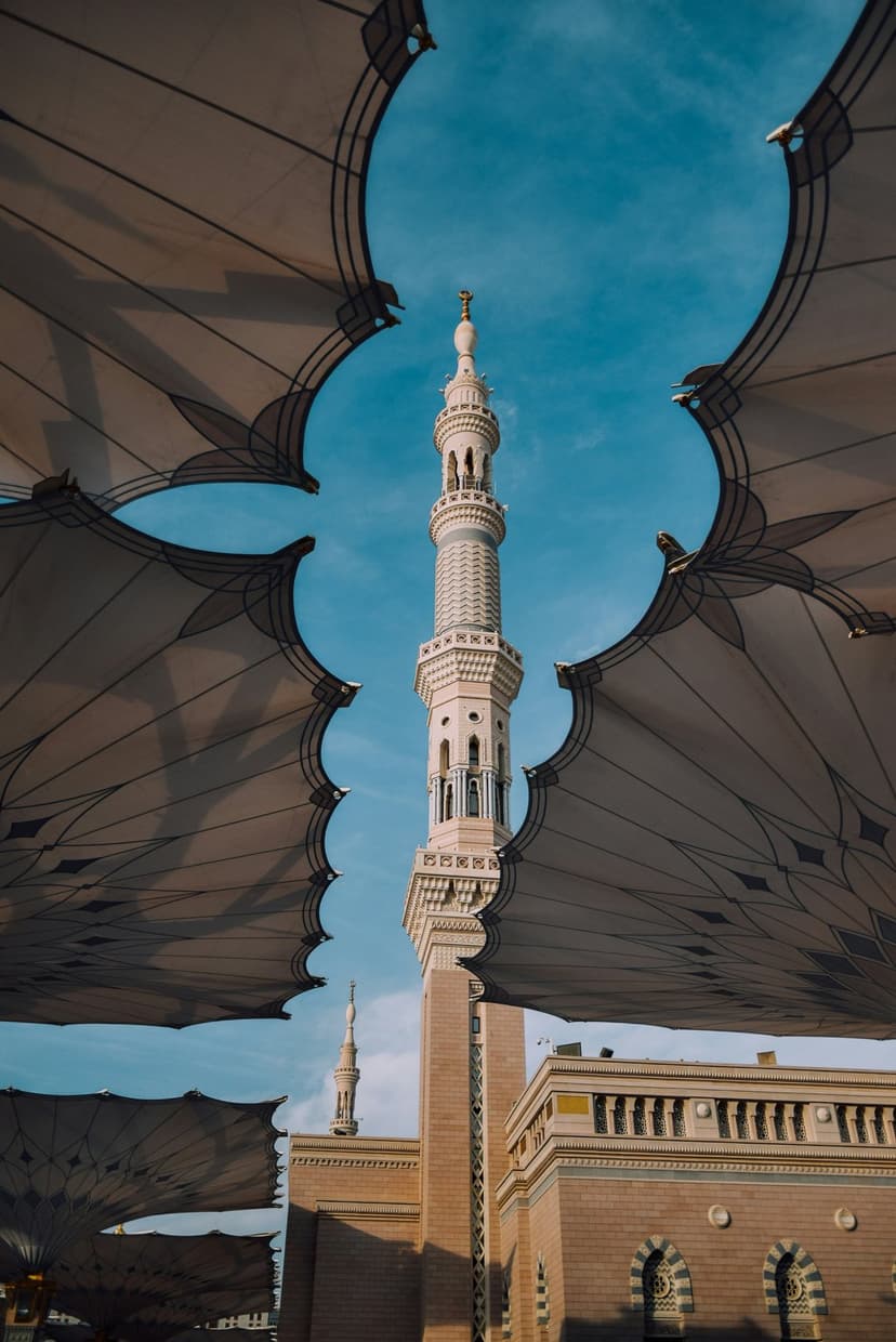 Intricate mosque minaret rises into a blue sky behind the geometric patterns of large, opened shade umbrellas.