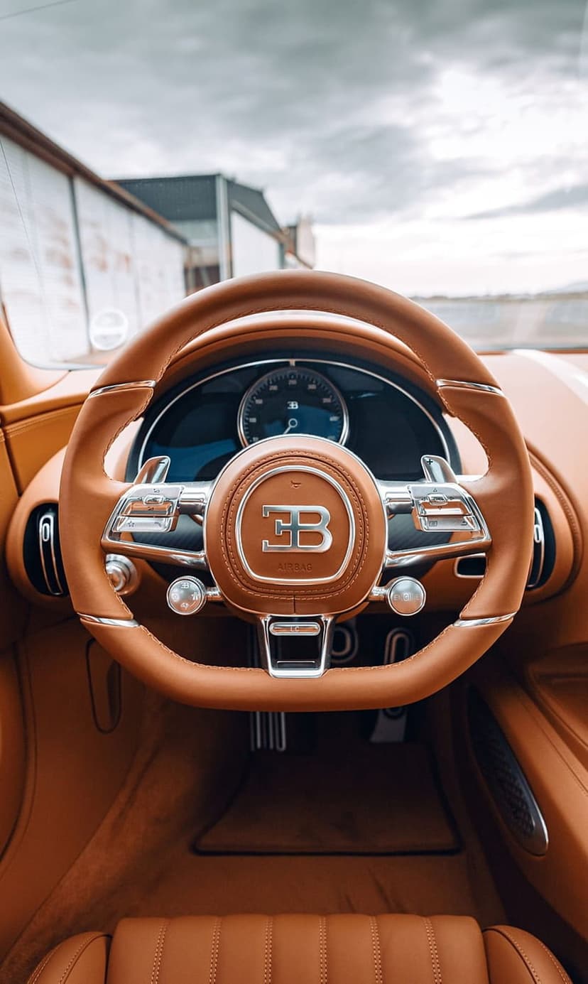 Bugatti steering wheel with tan leather and a chrome EB logo inside a luxury hypercar interior with pedals.