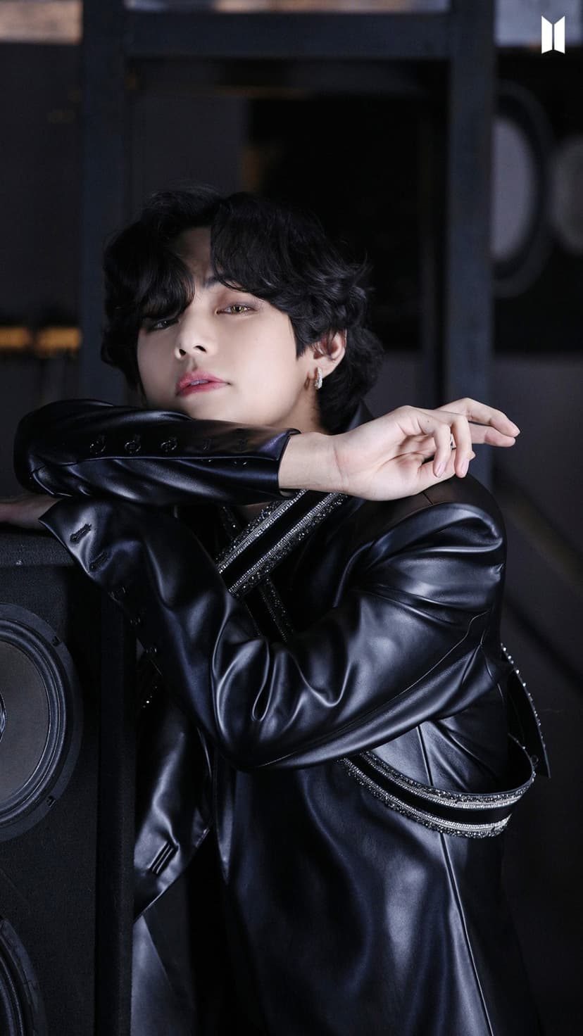 Kim Taehyung posing in a black leather jacket against a dark background with musical equipment in the distance.