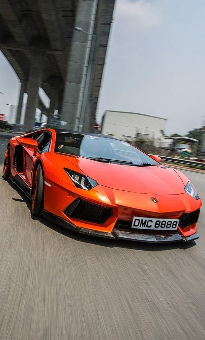 Orange Lamborghini Aventador speeds under a concrete bridge with motion blur and sharp headlights visible.