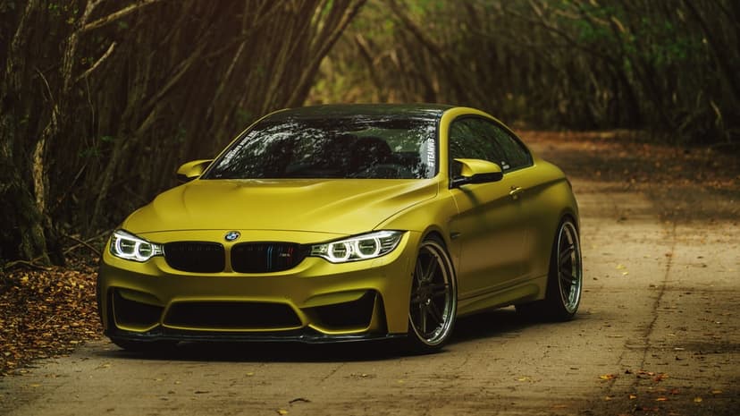 Matte yellow BMW M4 sports coupe with custom wheels on a paved forest road under a canopy of arching trees.