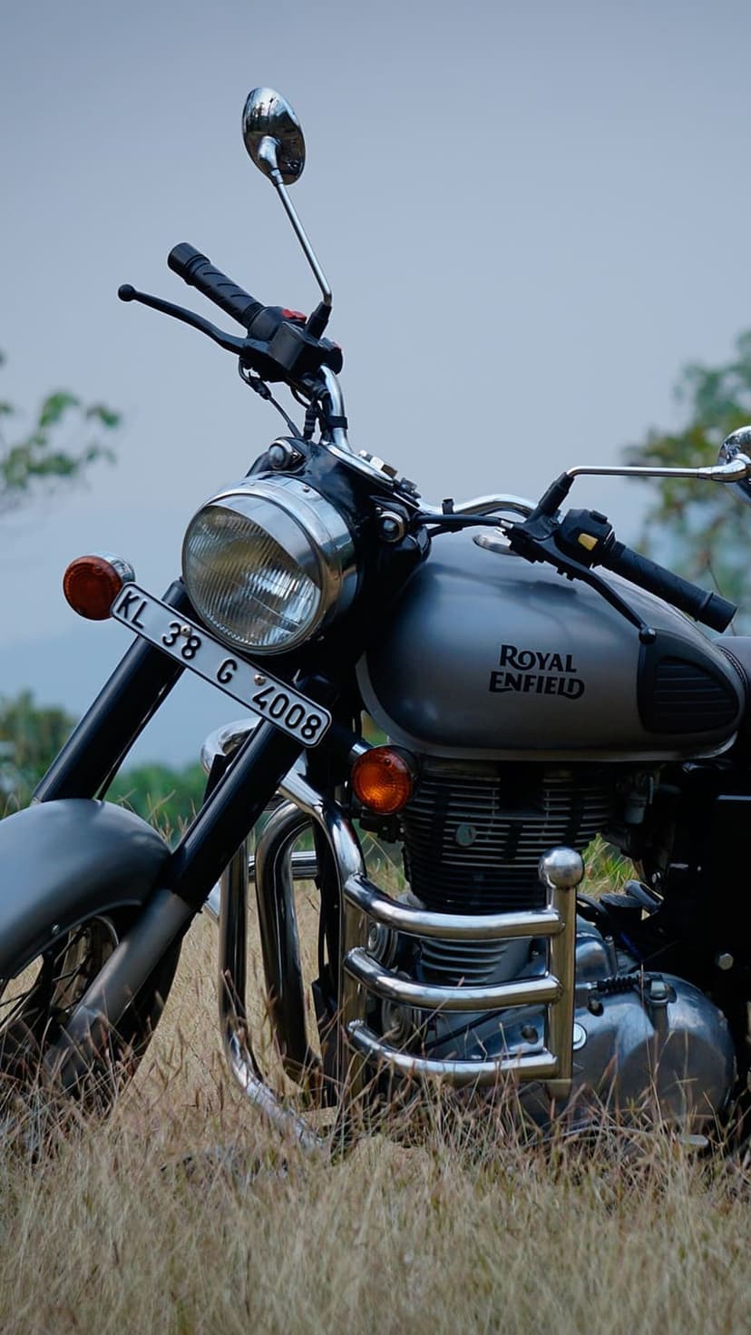 Silver Royal Enfield motorcycle with a chrome engine guard parked in a field of tall grass under a cloudy sky.