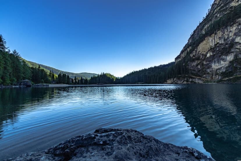 Sun-drenched mountain peak reflecting in a calm alpine lake with evergreen trees and a foreground rock at sunrise.