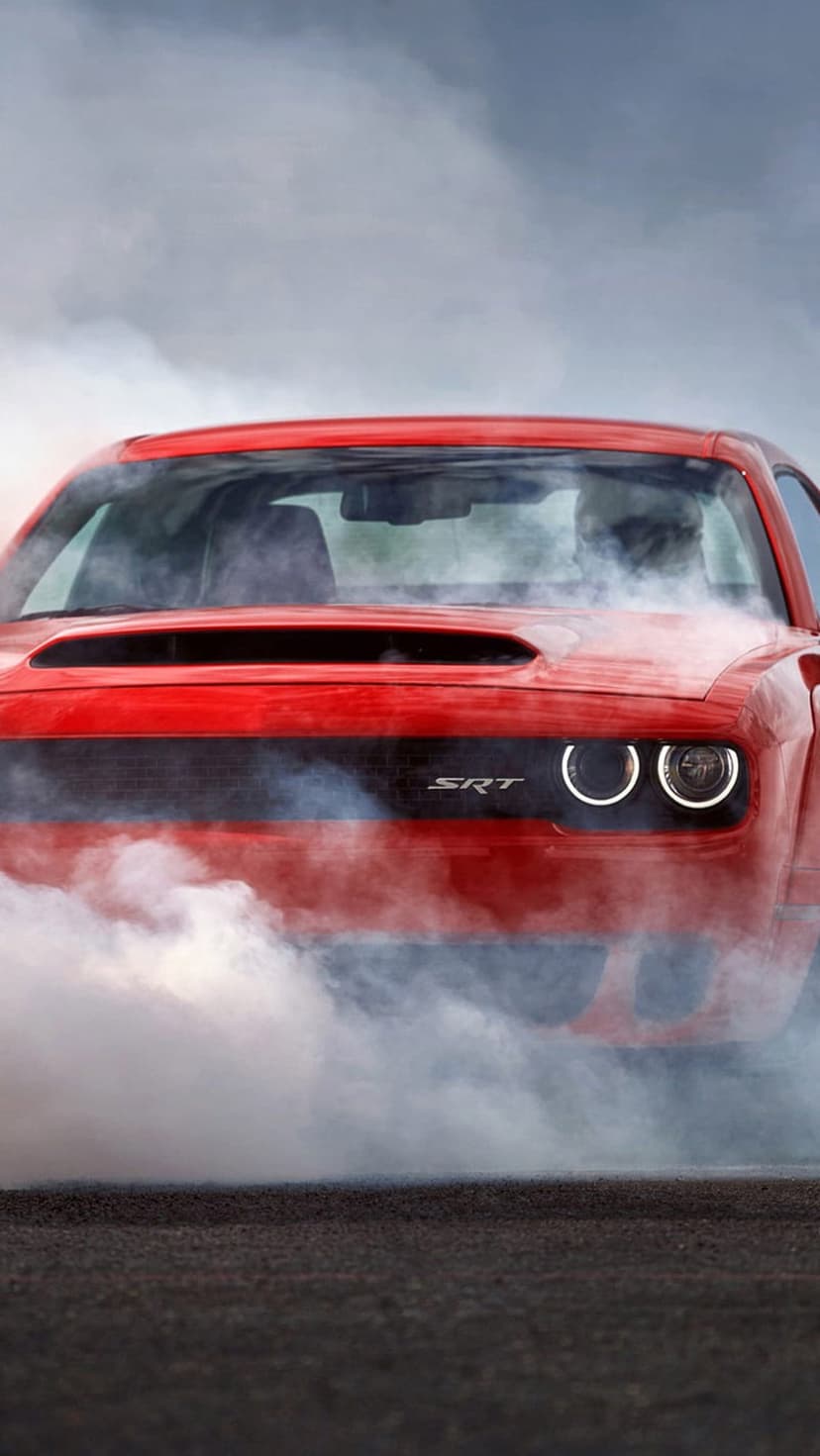 Red Dodge Hellcat muscle car emitting thick white tire smoke with glowing circular headlights and SRT grill logo.