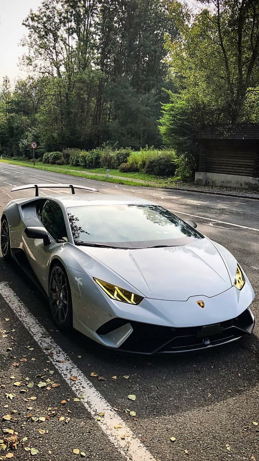 White Lamborghini Huracan with glowing yellow headlights parked on a rural road covered in fallen autumn leaves.