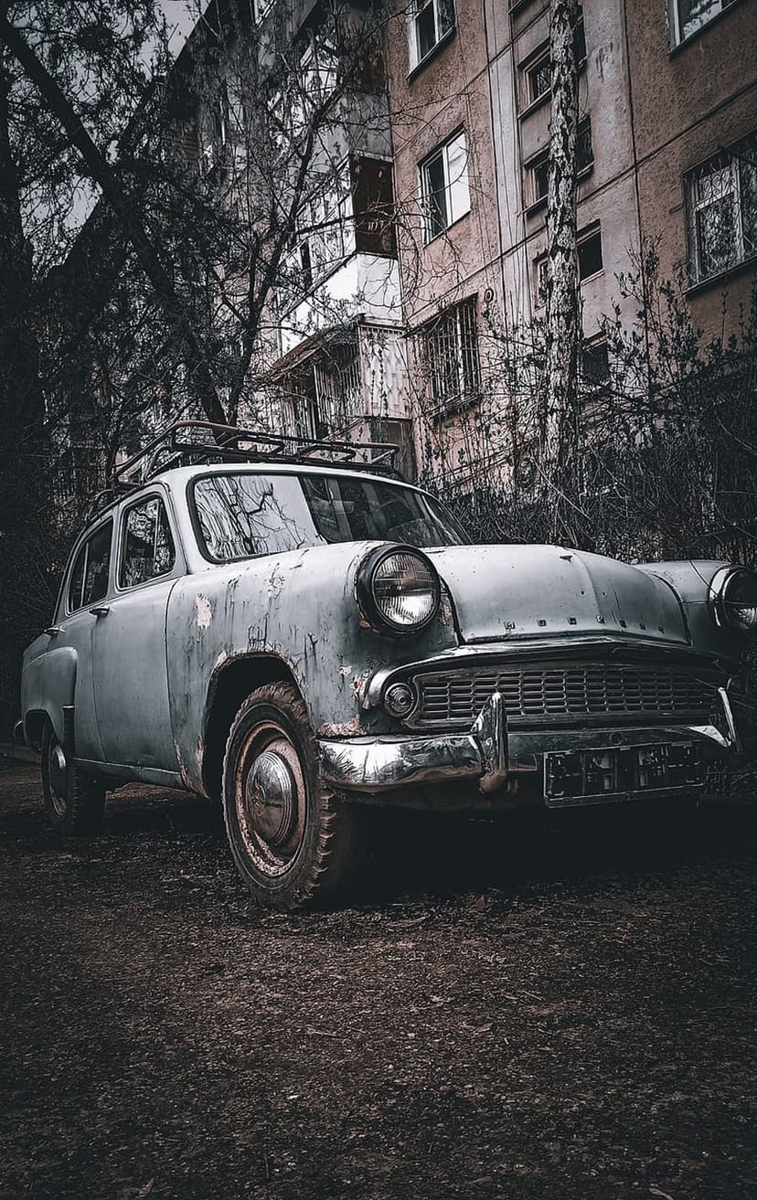 Rusted vintage car sits before a crumbling apartment building covered in thick vines on a dark gravel surface.