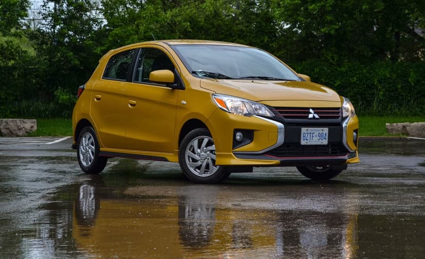 Yellow Mitsubishi Mirage hatchback parked on wet asphalt with reflections and a green tree background.